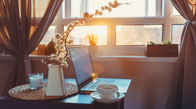 Home Work Place With Laptop, Cup Of Hot Drink And Blooming Brunch In Vase On Coffee Table Near Window On Sunset Or Sunrise. Freelance, Working From Home, Online Learning, Home Office. Slow Living